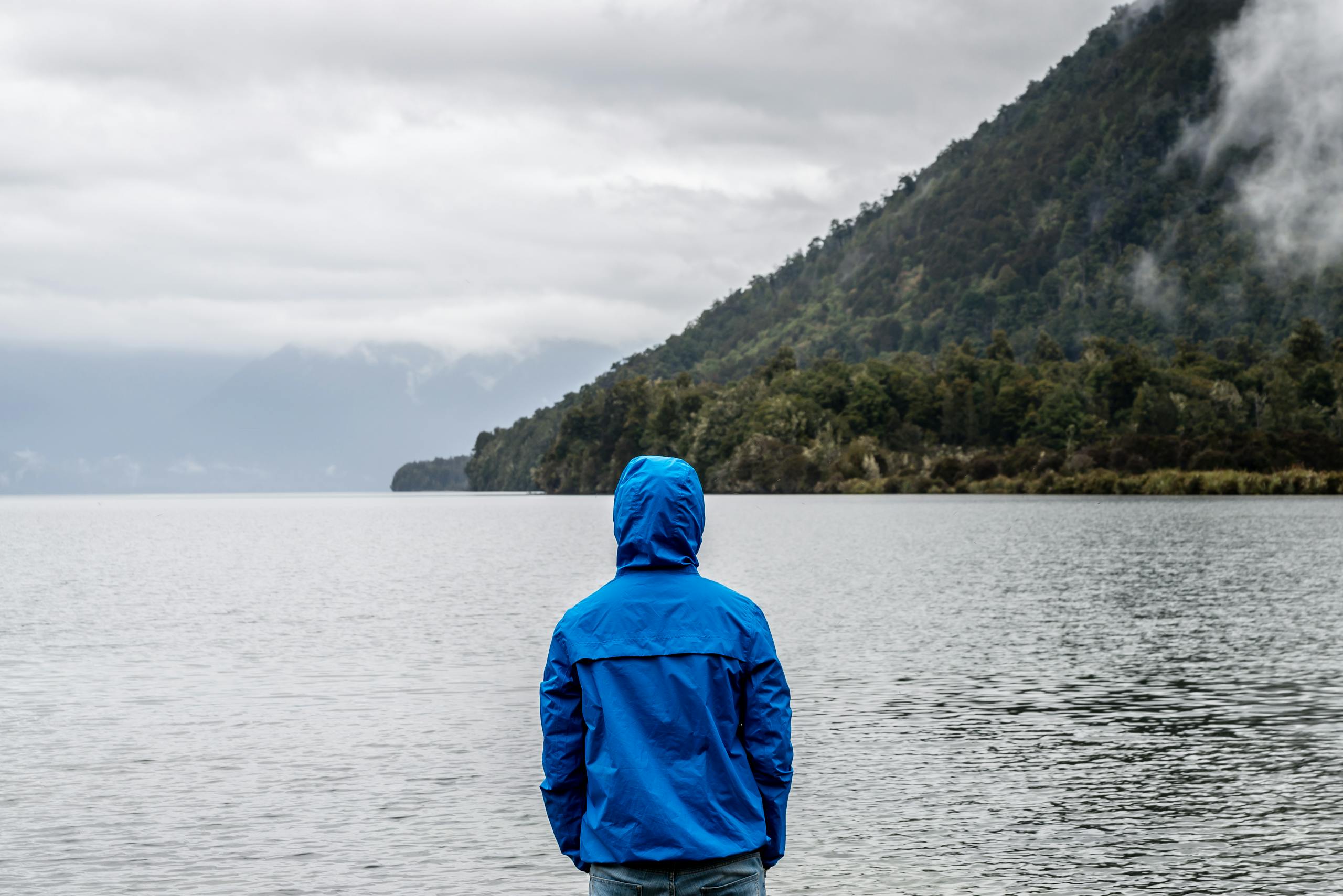 A person in a blue jacket stands by a misty New Zealand lake, surrounded by mountains.